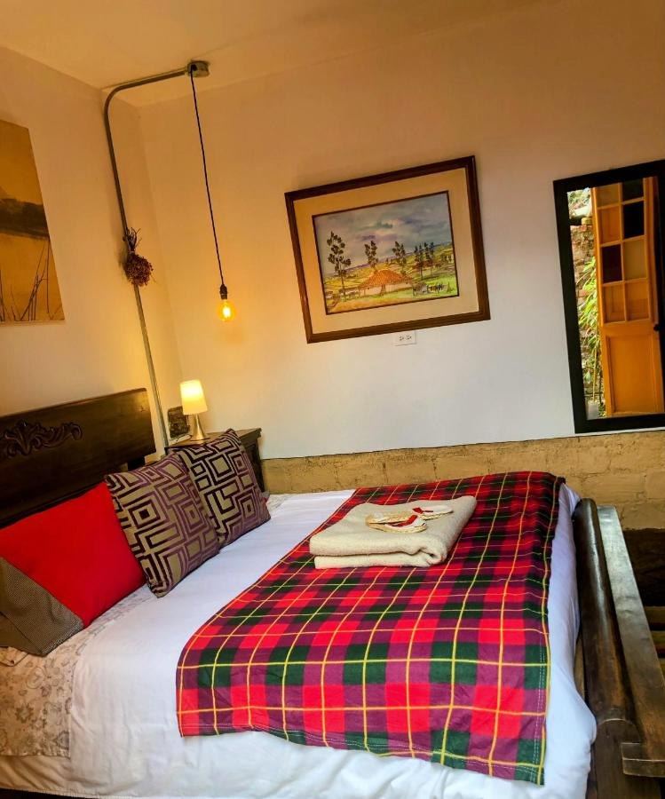Habitación La Chiquinquireña: cama Queen con manta escocesa bajo un cuadro de paisaje