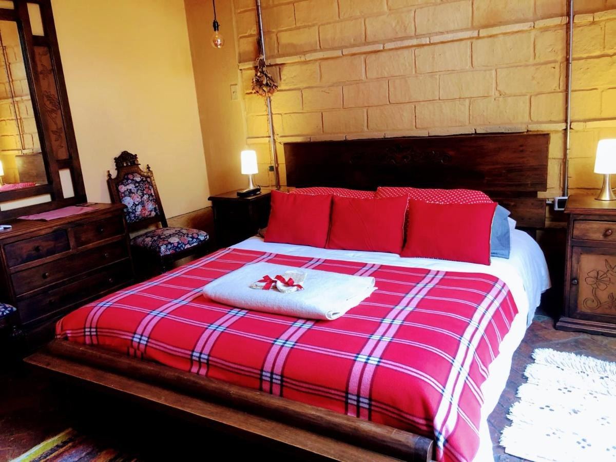 Habitación La Rústica: cama Queen con manta roja de cuadros y muro de piedra