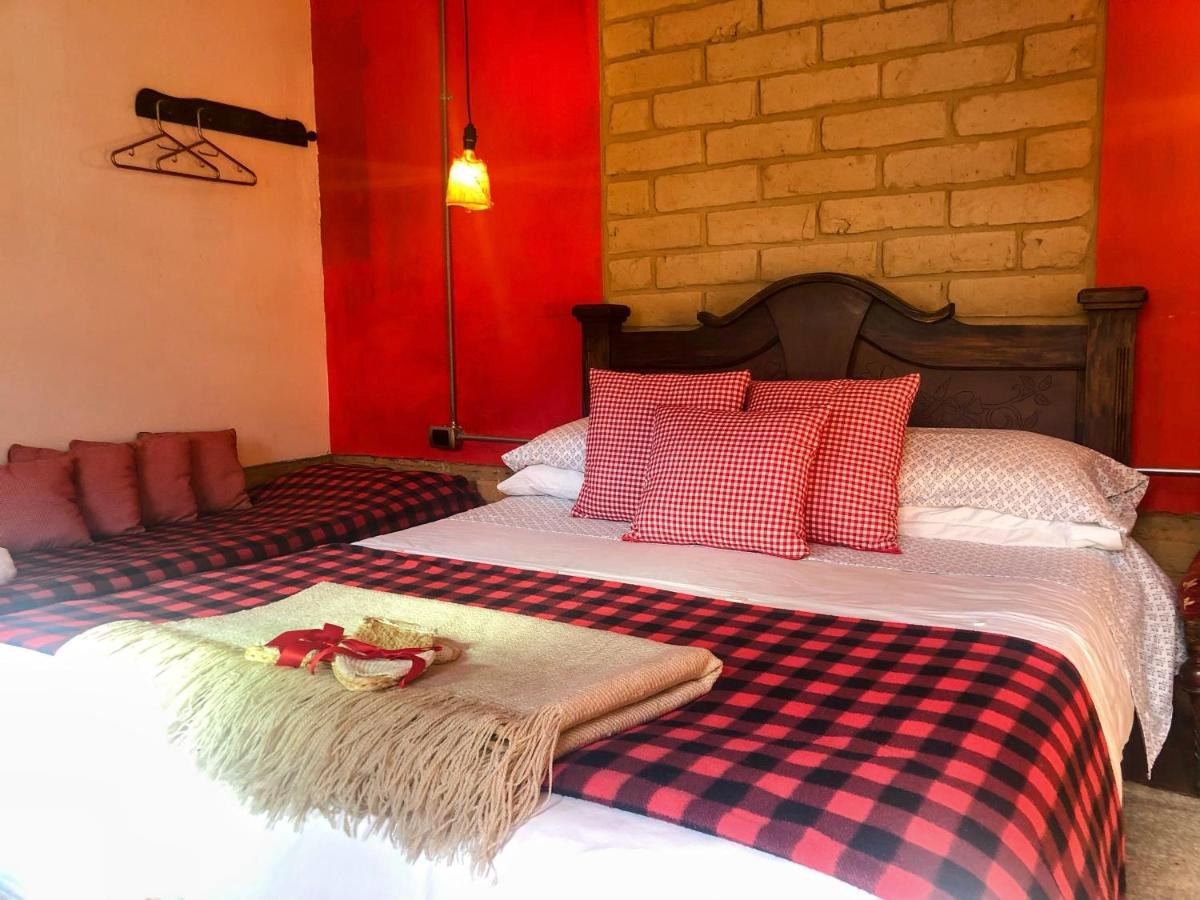 Habitación Los Vitrales: cama con manta de cuadros rojos y negros, pared roja y muro de adobe